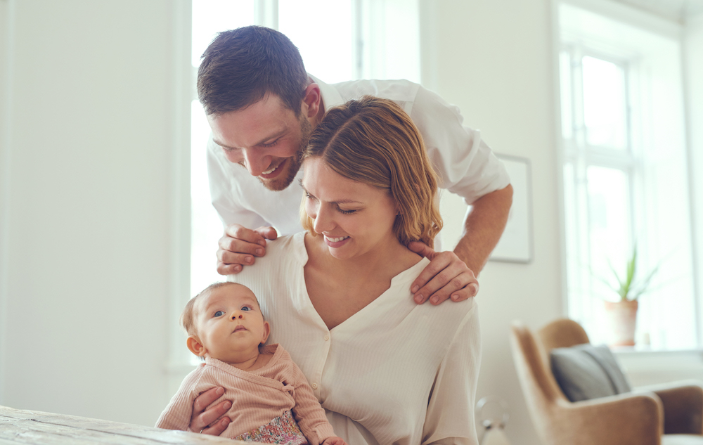 Smiling young couple sitting together at a table at home cradling their newborn baby daughter